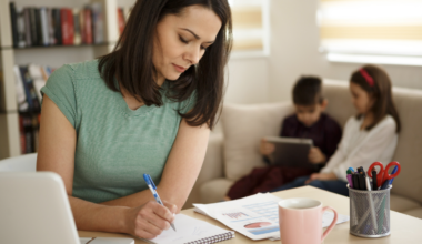 Mother writing things down her to-do list on a piece of paper with her children in the background. This depicts the invisible mental load of motherhood.